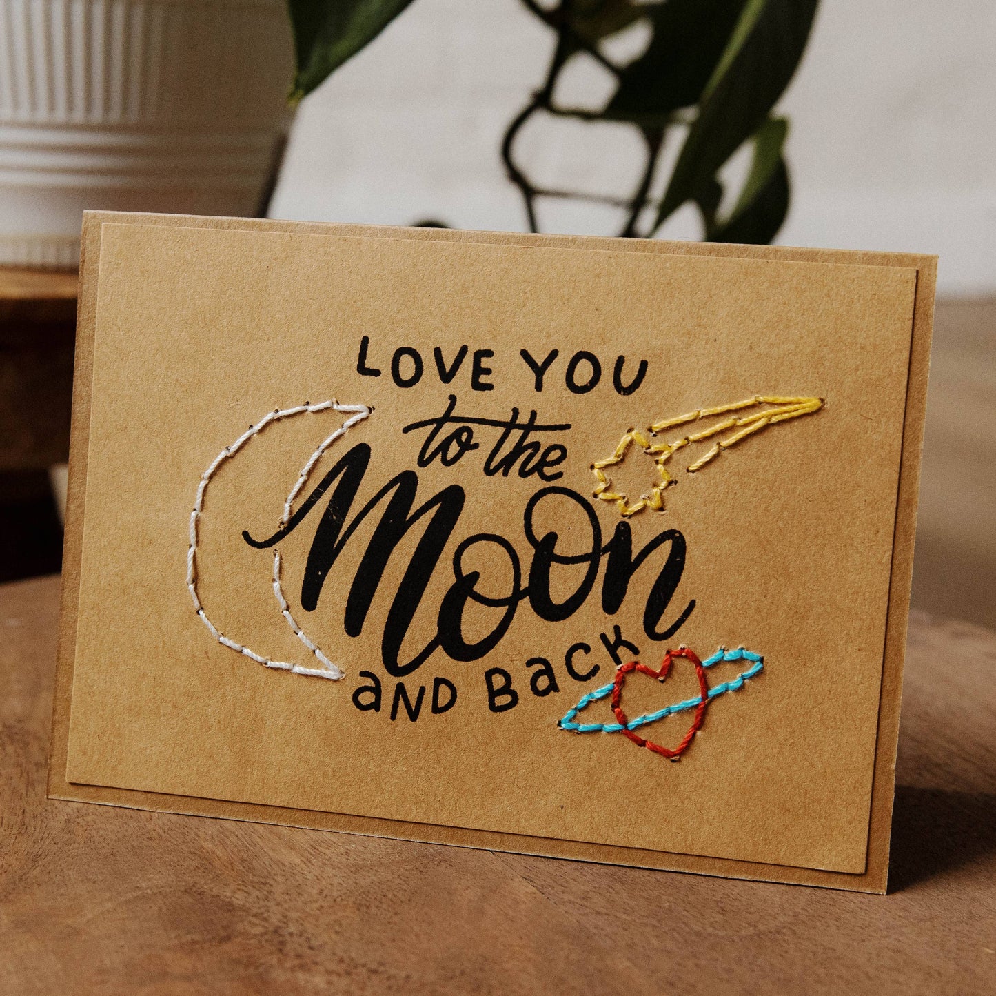 Card with embroidered text 'Love you to the moon and back' on a wooden surface.