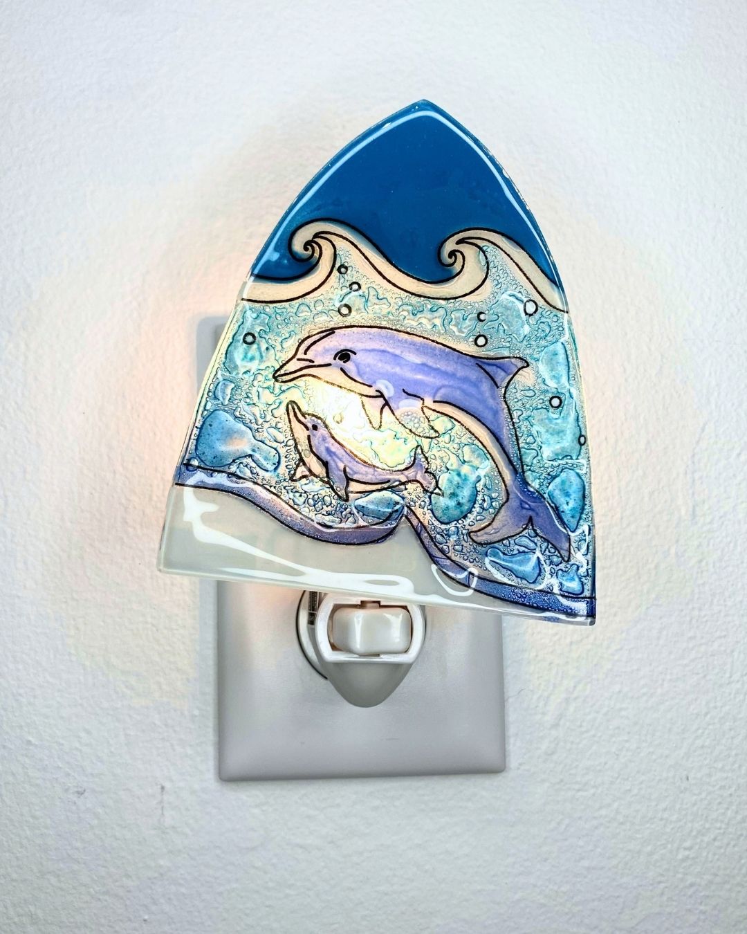 A glass night light shaped like a wave, featuring two dolphins swimming in blue ocean waves. The night light is shown plugged into a white outlet against a white textured wall, emitting a warm glow.