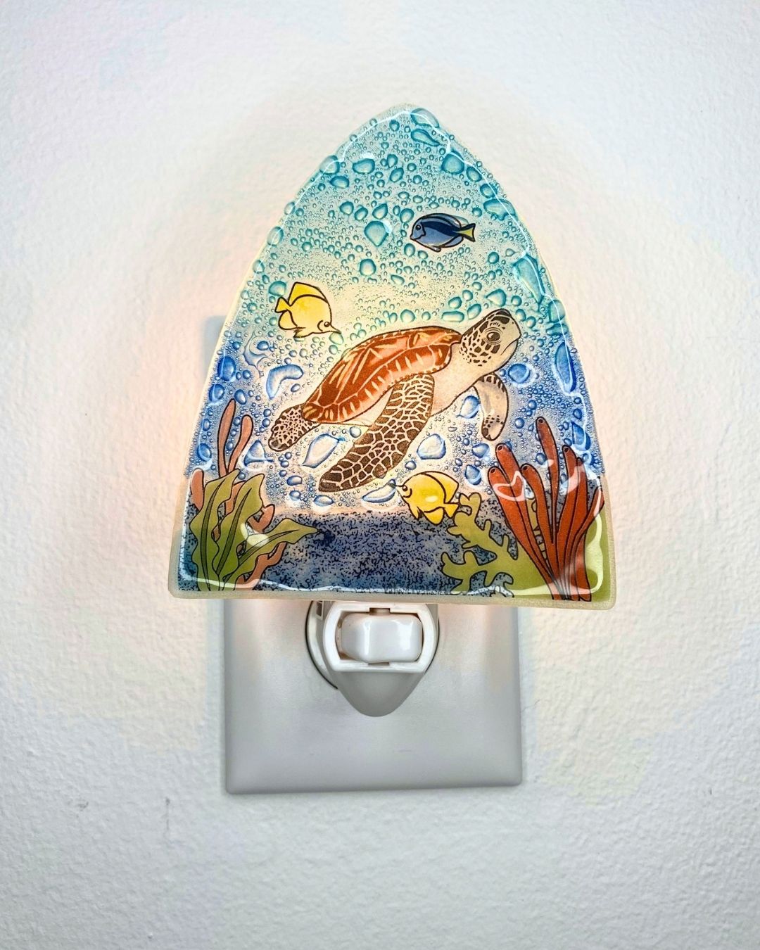 A decorative glass night light with a underwater scene mounted on a white wall outlet. The design shows a sea turtle swimming among blue and yellow fish, coral, and bubbles, with a warm light glowing from within.