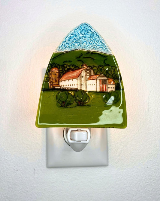 A lit triangular nightlight featuring a white barn against a green hill and flying the American flag is plugged into a white wall.