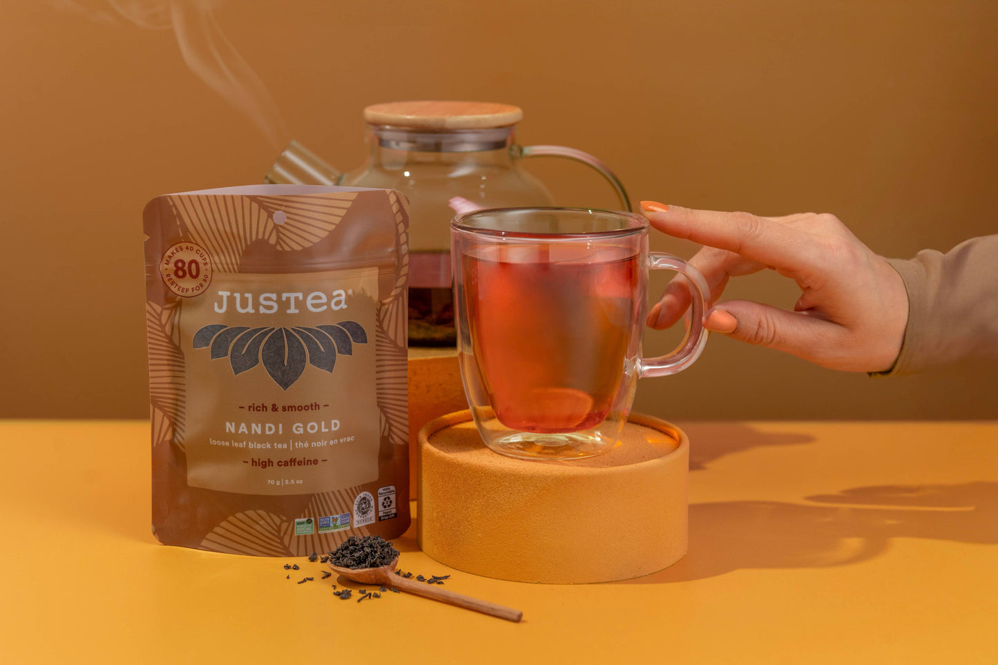 Hand holding a glass of tea next to a Justea tea package on a yellow surface.