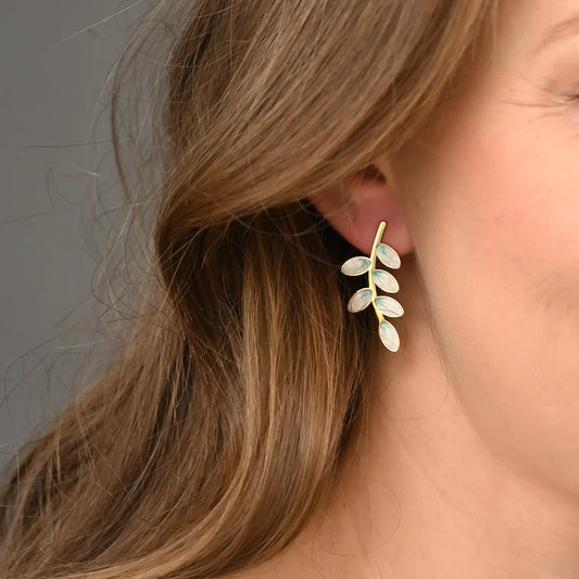 Close-up of a person wearing a gold and green leaf-shaped earring against a neutral background