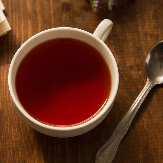A white ceramic cup filled with a deep red tea, placed on a dark wooden table next to a silver spoon.