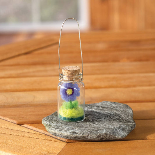 A small glass vial with a cork stopper and a thin metal loop handle, containing a felt flower arrangement in purple, green, and yellow, displayed on a gray stone slice over a wooden surface.