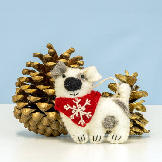A small white felt dog ornament with gray patches, a red bandana with a white snowflake, and a hanging loop, posed in front of two gold pinecones on a white surface against a light blue background.