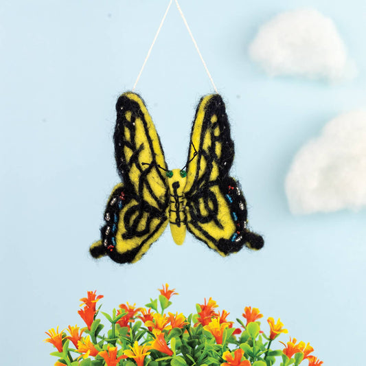 A yellow and black felted wool butterfly ornament hanging from a thin string against a light blue background, with decorative white clouds and small orange flowers at the bottom.