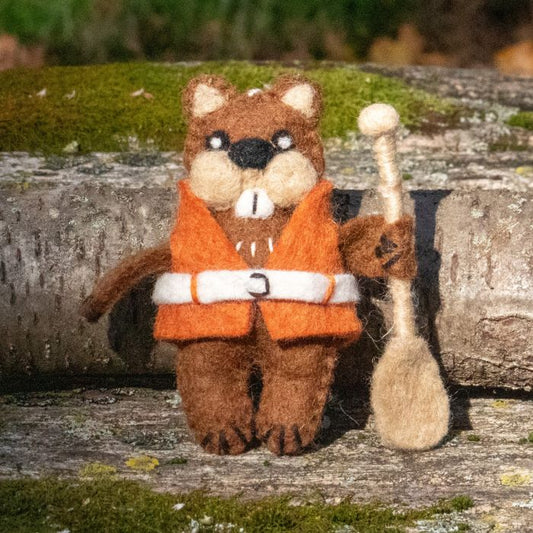 Brown beaver ornament wearing an orange lifejacket with white belt and holding a tan paddle set against a mossy log in an outdoor setting.