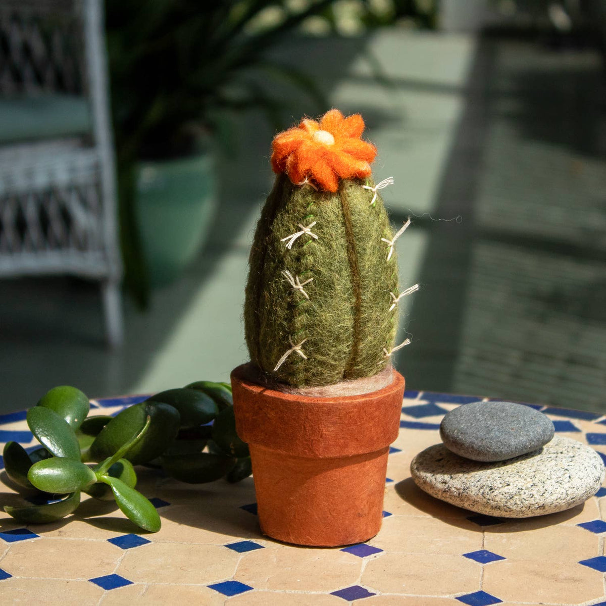 A felted wool cactus decoration in a terracotta pot, styled on a blue-tiled surface with a small succulent and stones in a sunlit patio setting.