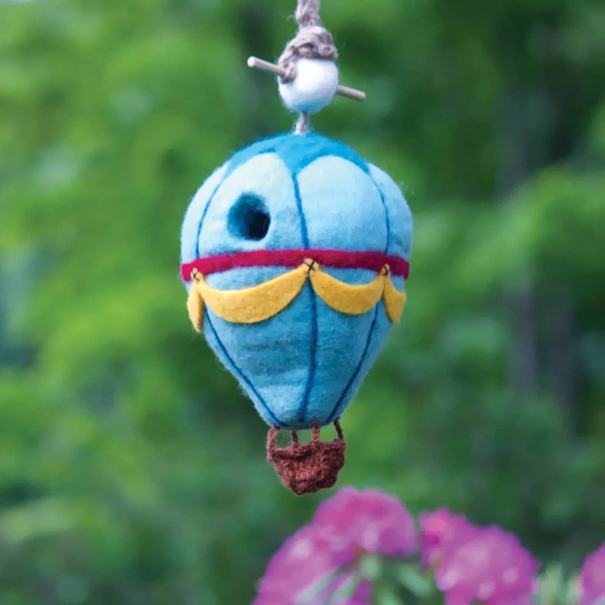 A small felt hot air balloon bird house hanging outdoors against a blurred green garden background with pink flowers in the foreground. The balloon is light blue with a red band, yellow scalloped trim, a brown basket, and a white bead with gray string at the top.