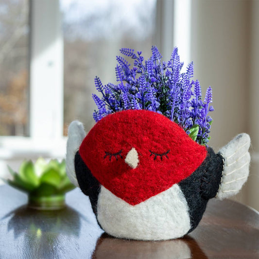 A woodpecker-shaped planter made from felt fabric, with a red face, black and white body, and white wings, holding a small cluster of purple artificial flowers. The planter sits on a dark wooden table in front of a softly blurred window.