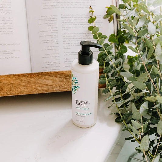 Thistle Farms eucalyptus mint hand lotion bottle on a surface with a plant and book in the background.