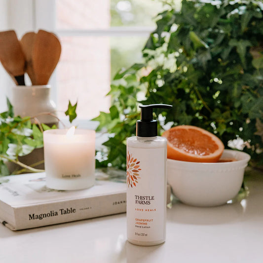 A pump bottle of grapefruit jasmine hand lotion is in a kitchen with a candle, cookbook, grapefruit, and utensils in the background.