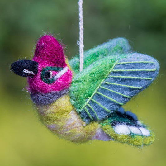 A handcrafted felt bird ornament with a bright pink head, green wings, yellow belly, and black beak, hanging by a white string against a soft blurred green background.