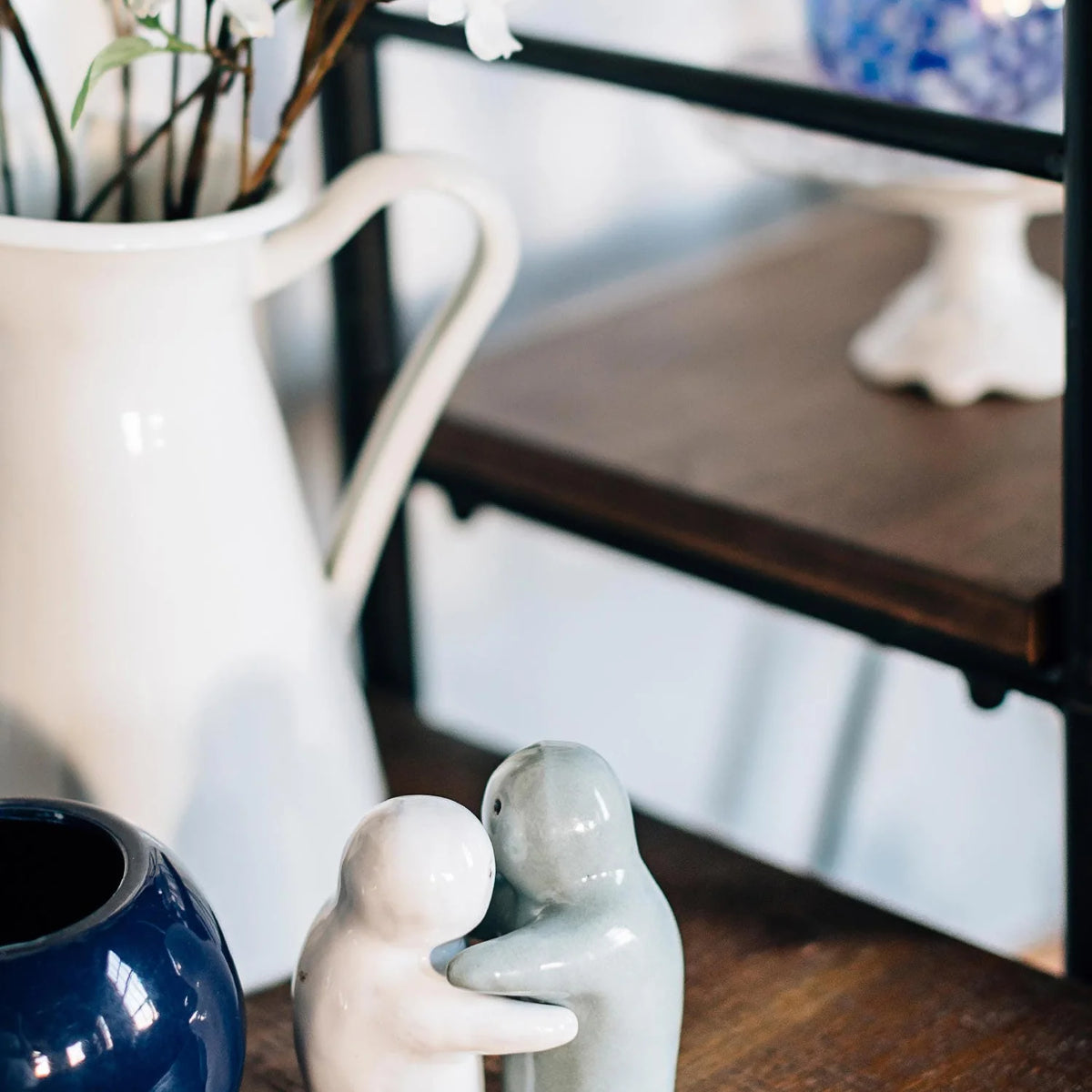 Two small glossy ceramic figurines of embracing figures, one in off-white and the other in pale blue-gray, displayed on a wooden shelf next to a white pitcher and a dark blue vase.