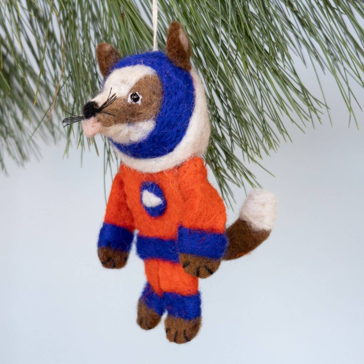 A felted wool dog ornament shaped like a fox wearing an astronaut suit, hanging from a pine branch against a pale blue background.