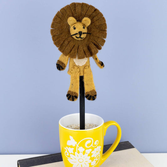 felted lion on a black stick next to a yellow mug with a floral design on a light blue background