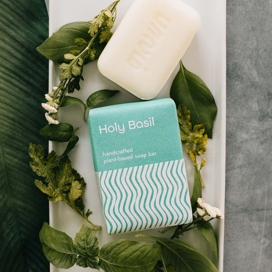 A white bar of soap and its teal and white rectangular package are laying on a white tray and surrounded by green leaves and small flowers.