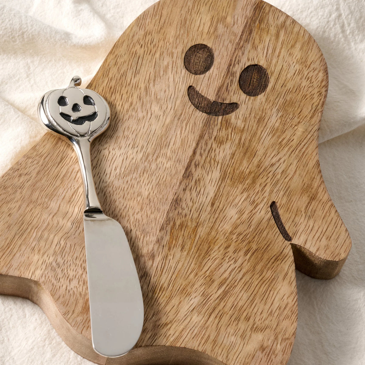 A silver metal cheese knife with a carved jack-o'-lantern handle rests on a wooden serving board shaped like a ghost, decorated with a simple face and set against a soft white fabric background.