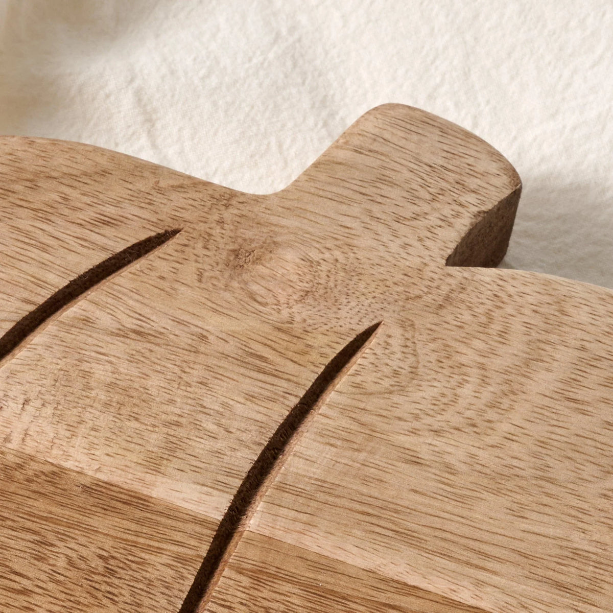 Close-up of a wooden serving board or tray with a natural oak grain, featuring two dark engraved diagonal lines, resting on a textured off-white surface.