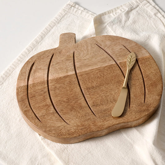 A pumpkin-shaped wooden serving board with carved leaf-like grooves and a small handle, styled on a white textured cloth with a gold-toned butter knife resting on top.