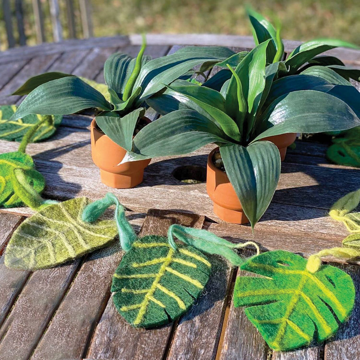 Green felt monstera leaf garlands with yellow-green vein details and hanging loops, displayed on a weathered wooden table alongside potted plants.