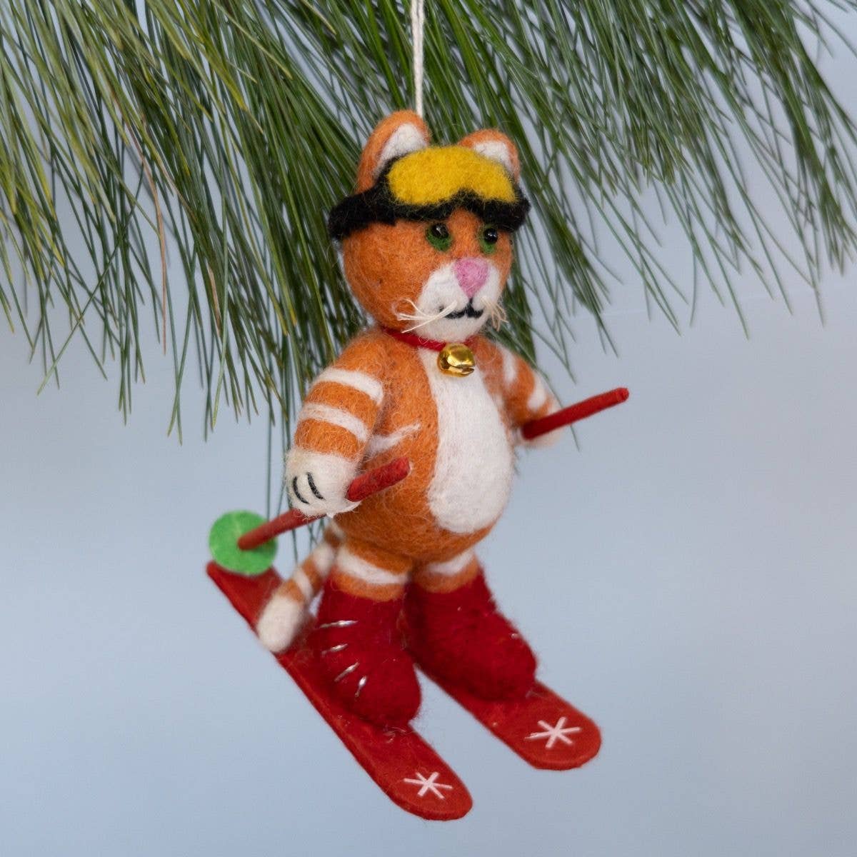 A felt cat Christmas ornament dressed as a skier, hanging from a pine tree branch against a light blue-gray background.