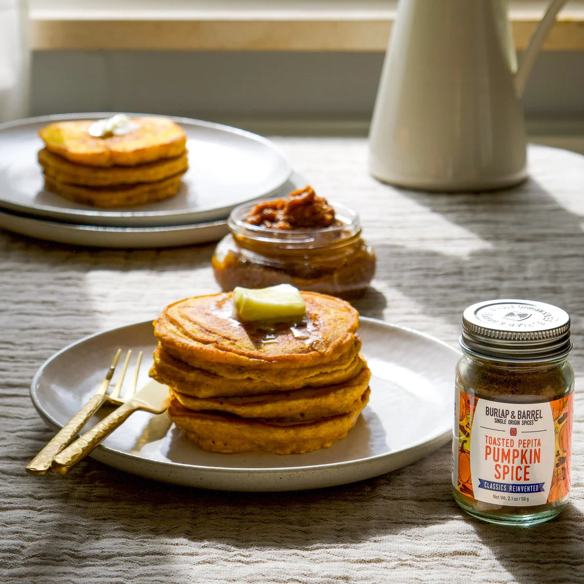 A plate of pumpkin spice pancakes sits next to a jar of toasted pepita pumpkin spice on a table.
