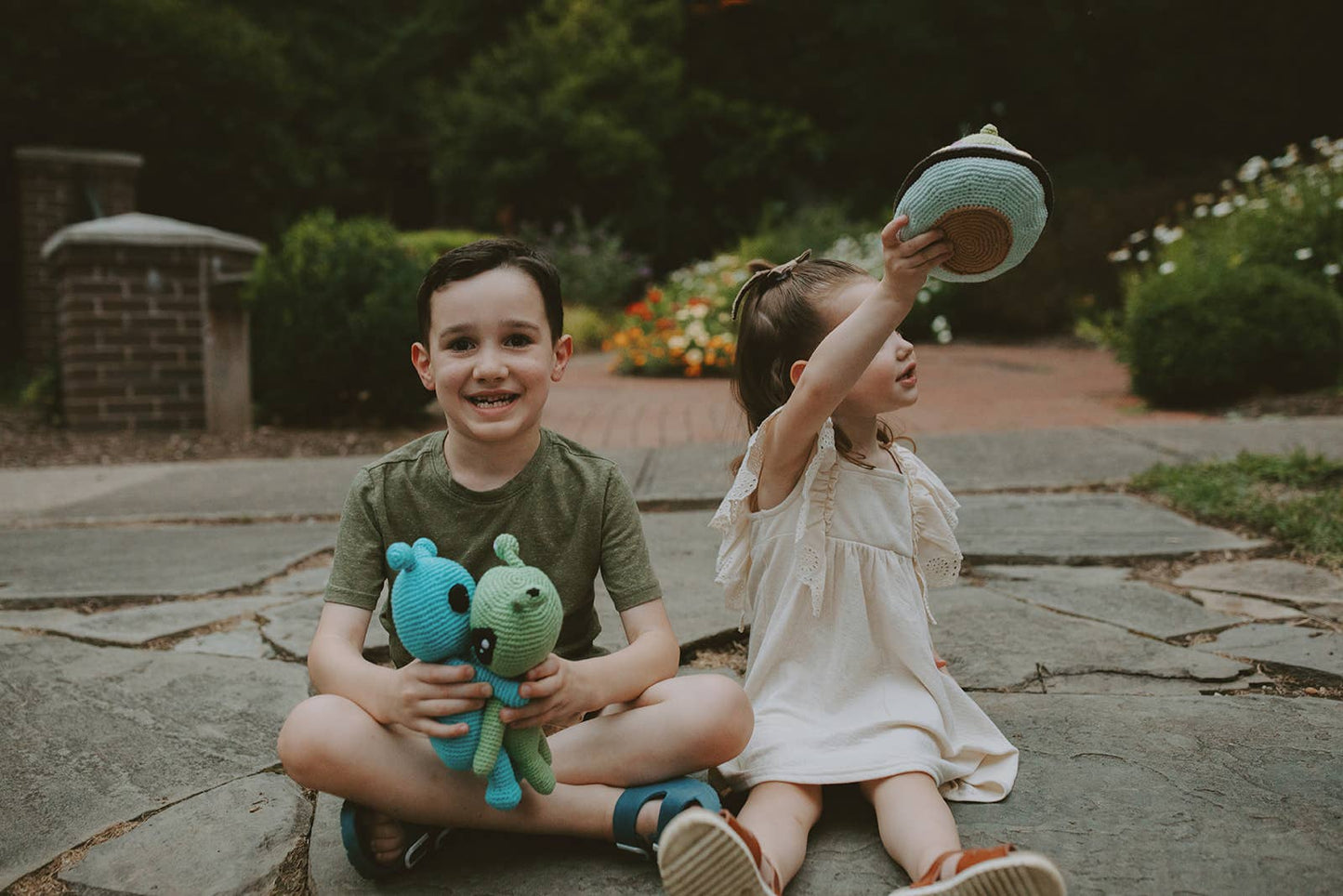 Two children sitting on a stone path outdoors, one holding two alien toys and the other making a stuffed UFO fly.