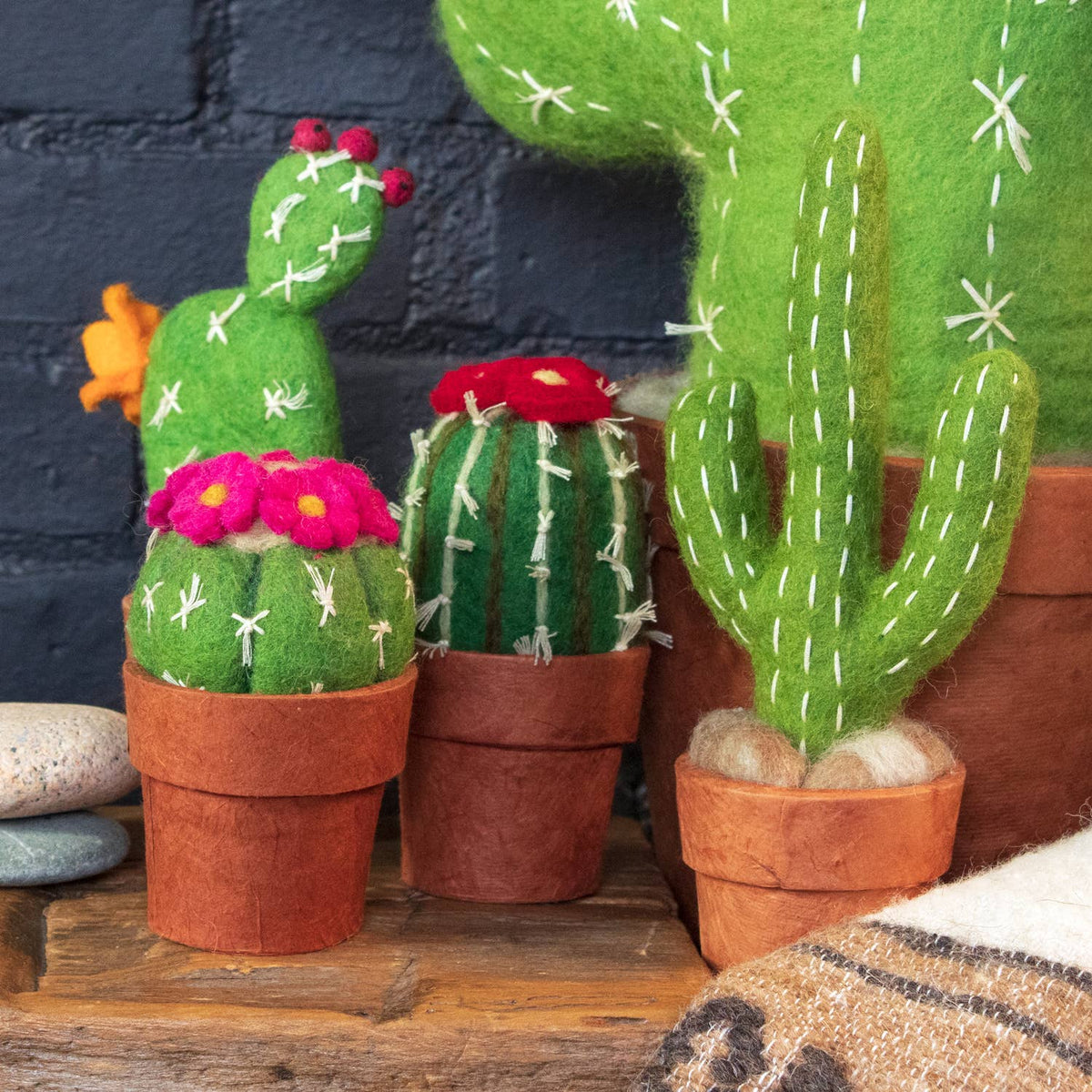 A set of felt cactus and succulent decorations in terracotta pots, displayed on a wooden surface against a dark brick wall. The collection includes various green felt cacti with white stitched details, some adorned with red and pink felt flowers.