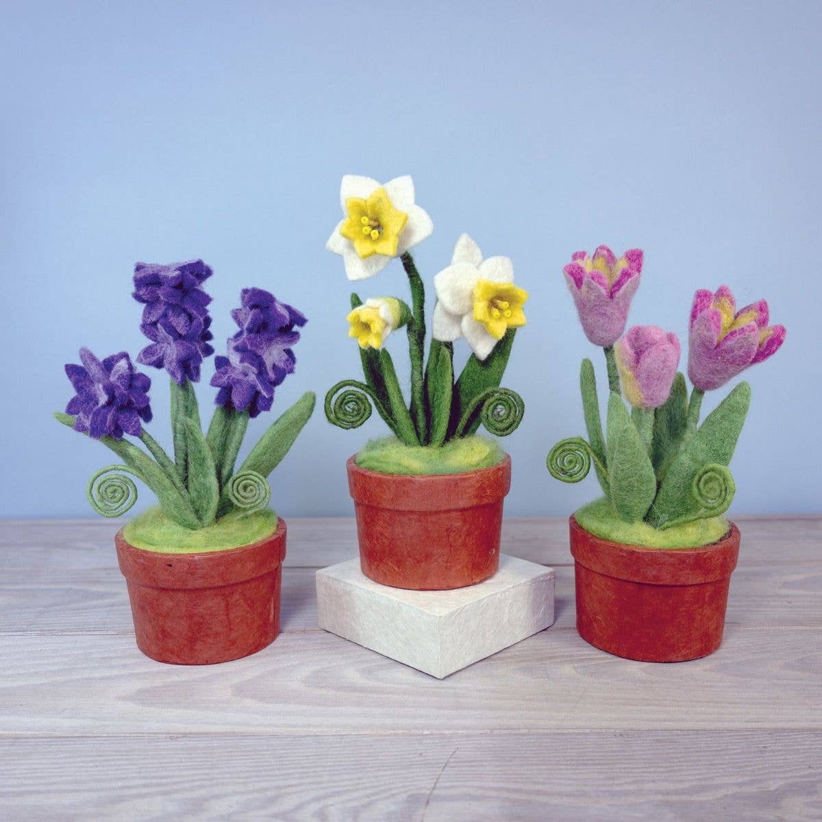 Three felted flower pots with flowers on a wooden surface and light blue background