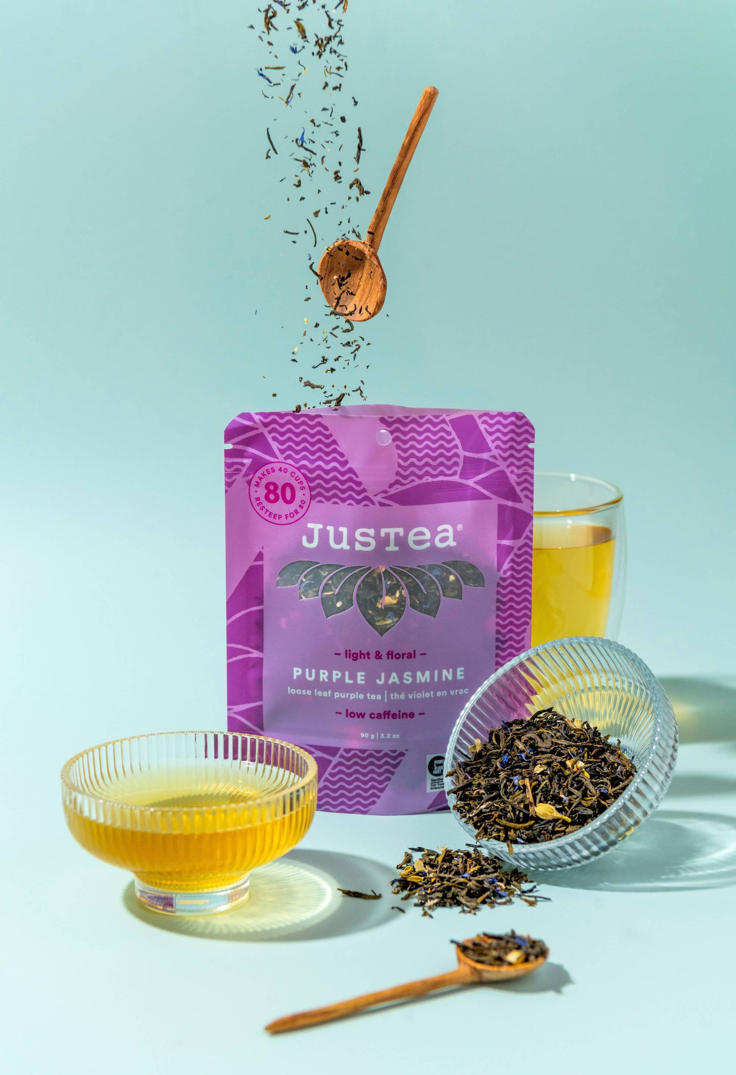 Justea Purple Jasmine tea packaging with a glass of tea and tea leaves on a light blue background