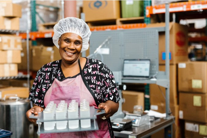 A smiling woman in a white hairnet and pink apron holds a tray of small white bottles in a warehouse or production setting with shelves, boxes, and equipment in the background.