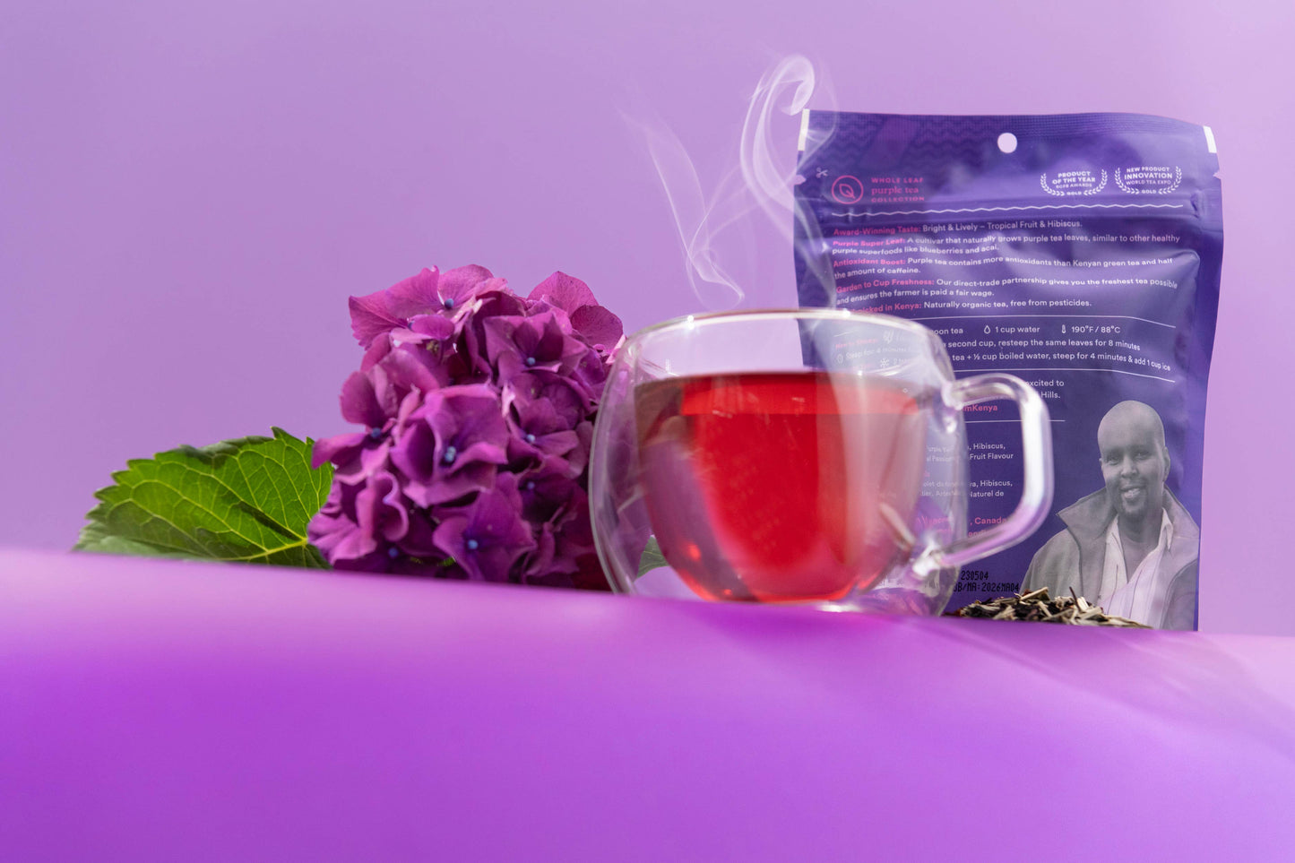 Tea bag with a purple package featuring a person, a glass of red tea, and purple flowers on a purple background.