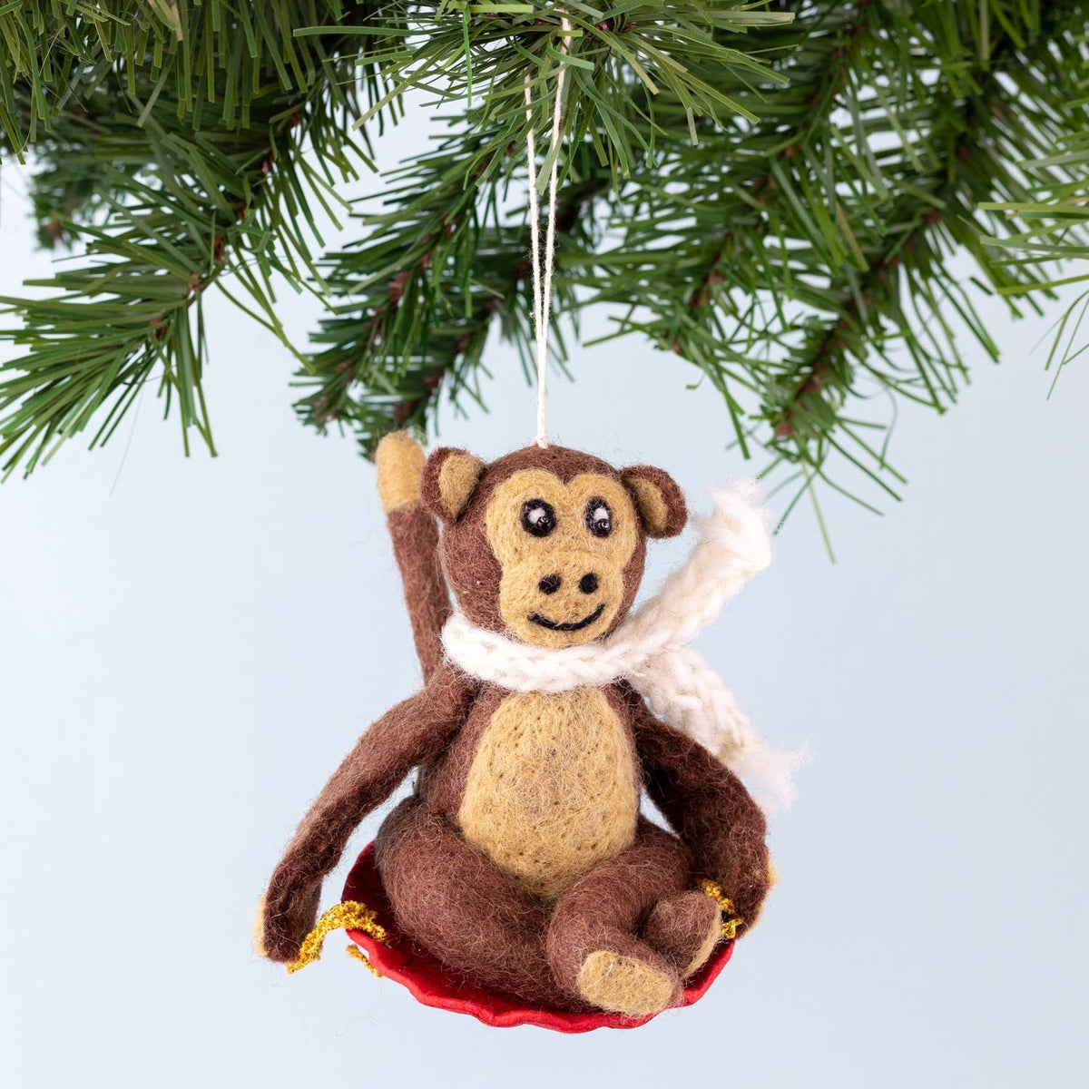 A felt monkey Christmas ornament hanging from a green pine tree branch against a light blue background. The ornament is brown and beige with a white knit scarf and a red seat.