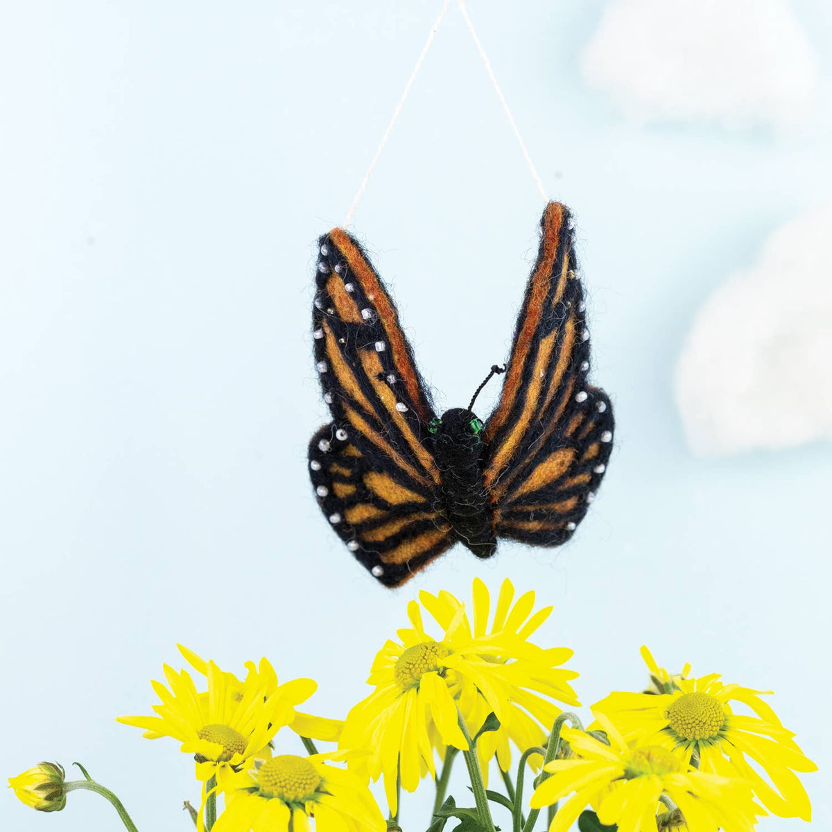 A felt butterfly ornament with orange, black, and white striped wings hanging from a thin string above a cluster of yellow flowers against a pale blue background.