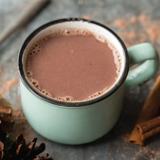 Teal mug filled with hot chocolate on a rustic surface with cinnamon sticks and pine cones.
