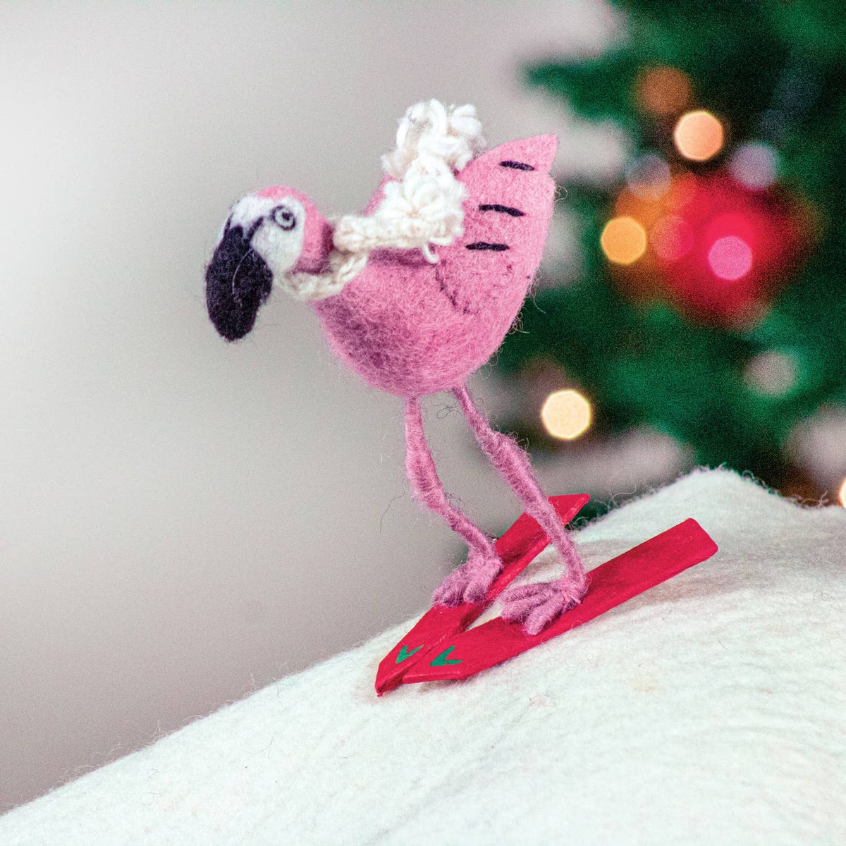 A pink felt flamingo figurine wearing a white knit scarf and red skis, posed on a white textured surface with a blurred Christmas tree in the background.