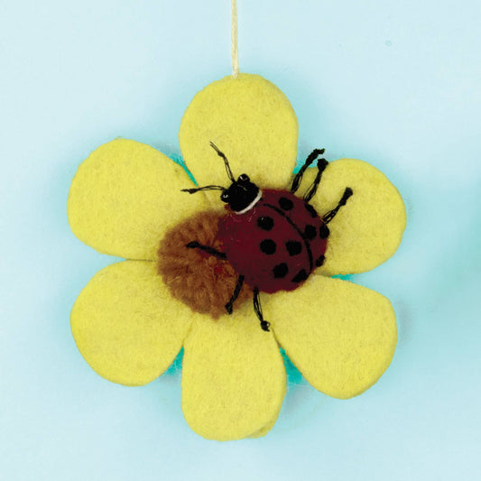 A handmade felt flower ornament with yellow petals, a brown fuzzy center, and a red ladybug figure on top, hanging against a light blue background.