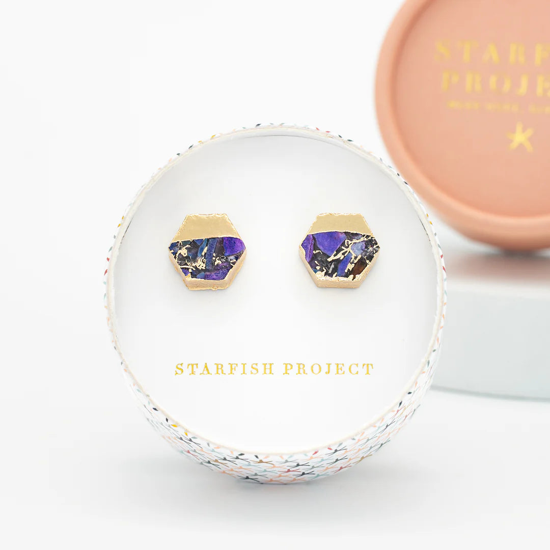 Hexagonal purple and gold stud earrings in a circular box.