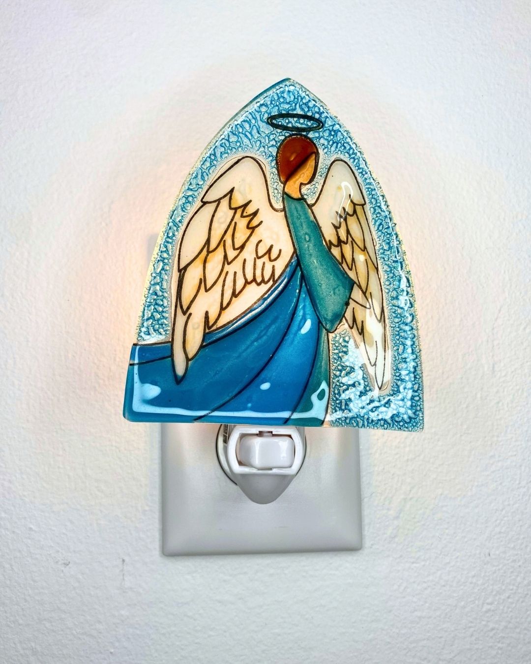 A lit triangular shaped nightlight featuring an angel with spread white wings and a blue robe is plugged into a wall outlet.