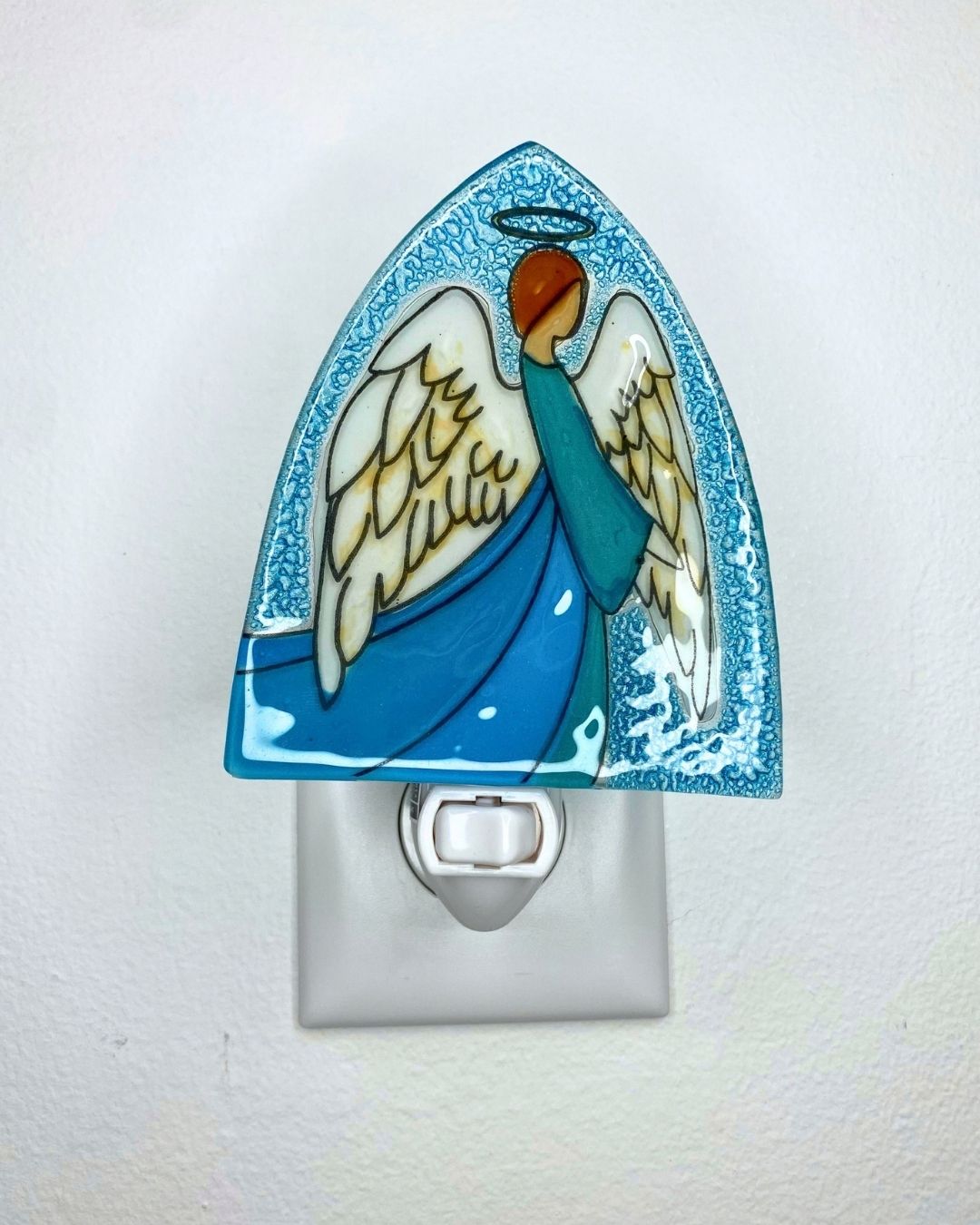 An unlit triangular shaped nightlight featuring an angel with spread white wings and a blue robe is plugged into a wall outlet.