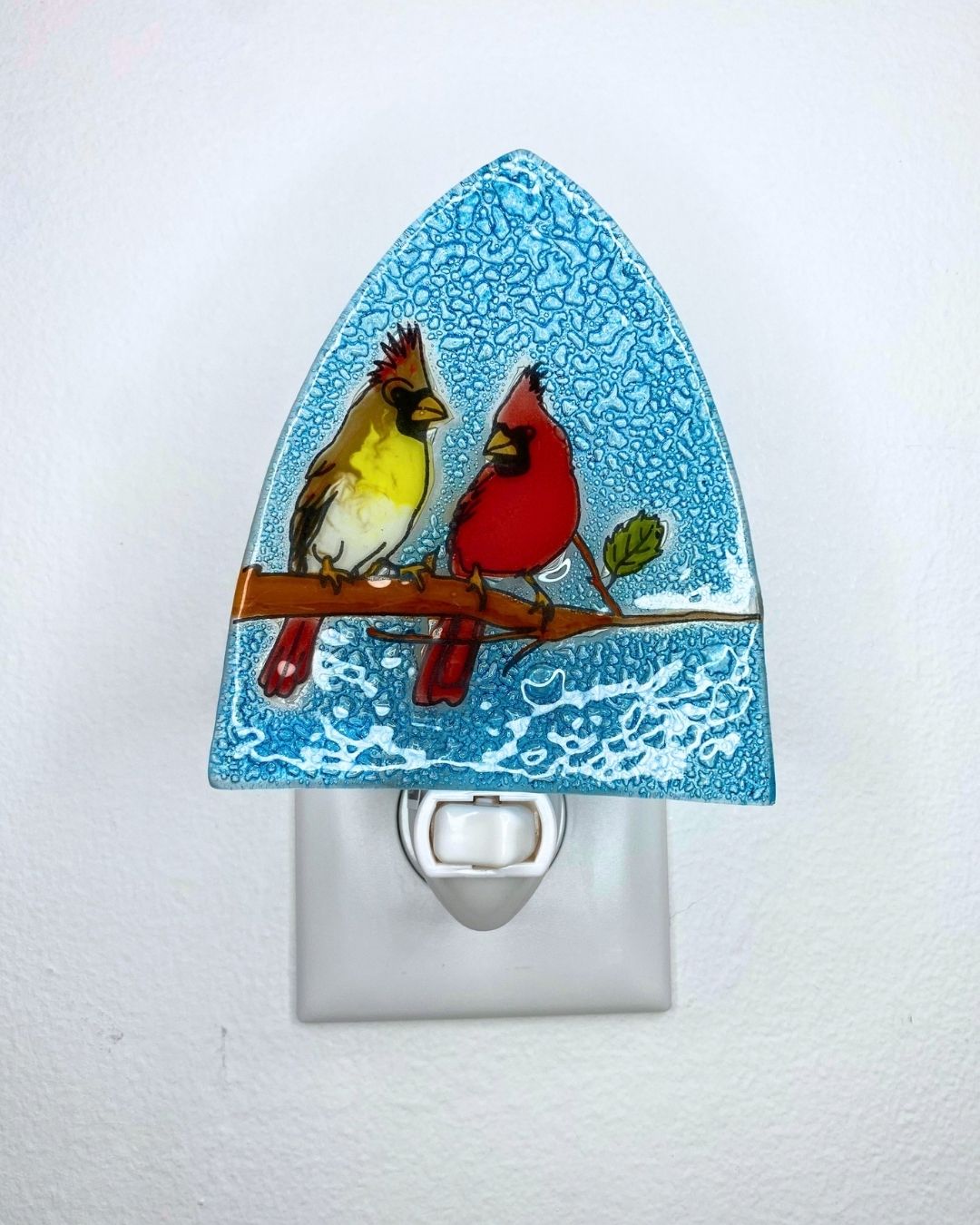 A decorative glass night light with an arched top and a white plug, featuring a male and female cardinal perched on a branch against a blue textured background.