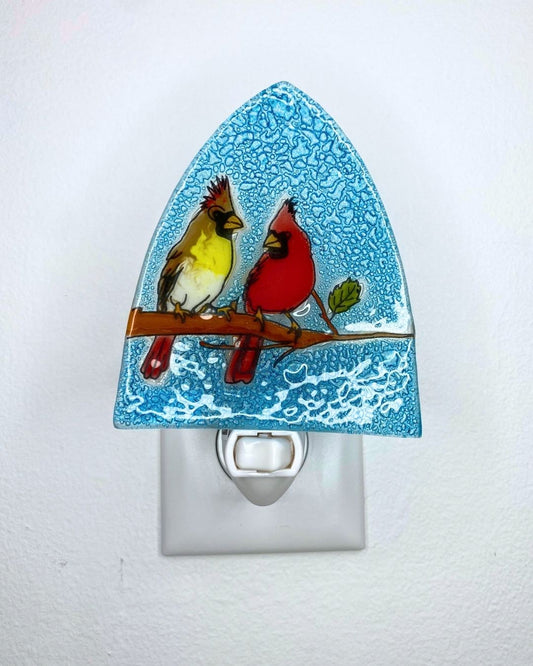 A decorative glass night light with an arched top and a white plug, featuring a male and female cardinal perched on a branch against a blue textured background.