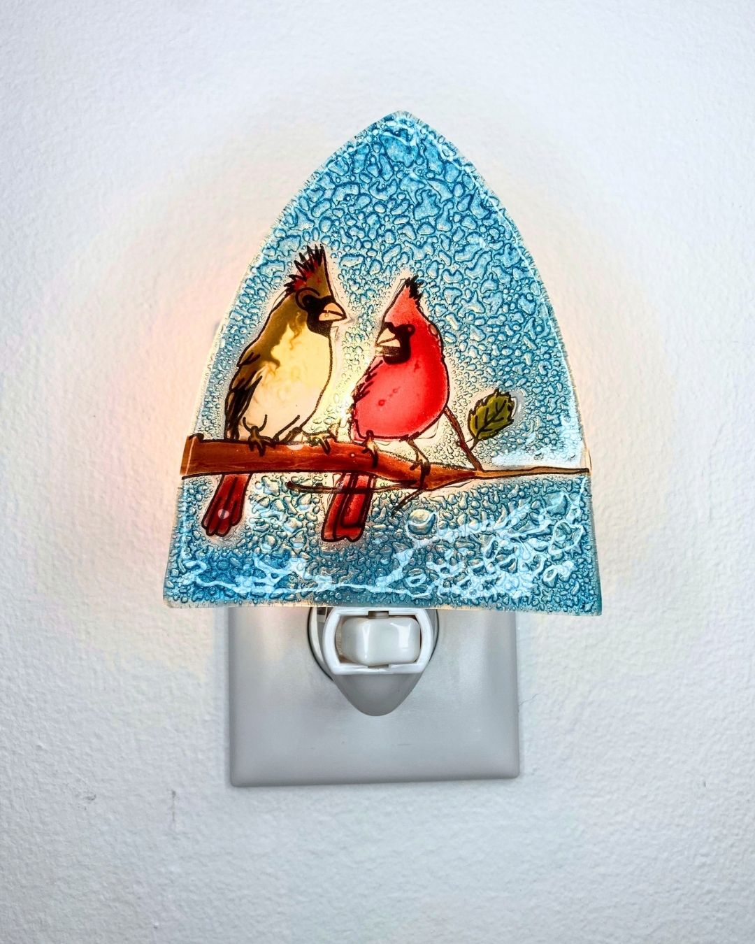 A lit up decorative triangular-shaped night light with two cardinal birds perched on a branch, shown plugged into a white wall outlet against a white wall.