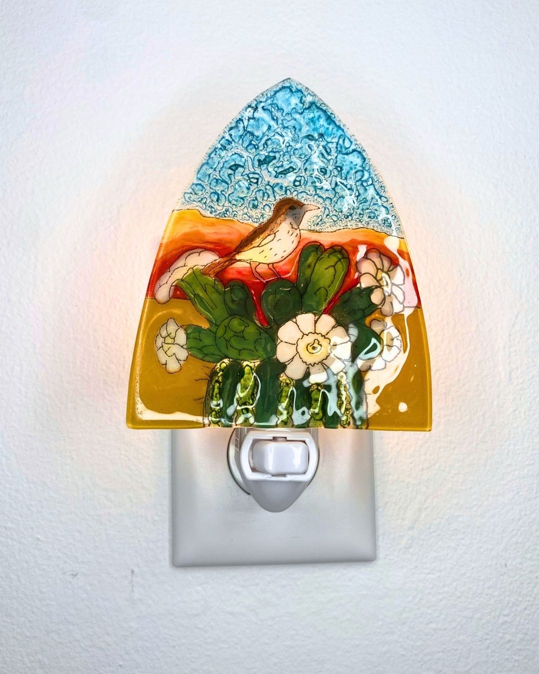 A lit decorative glass night light plug-in with a bird sitting on a green flowering cactus against a desert background, glowing against a plain white wall.