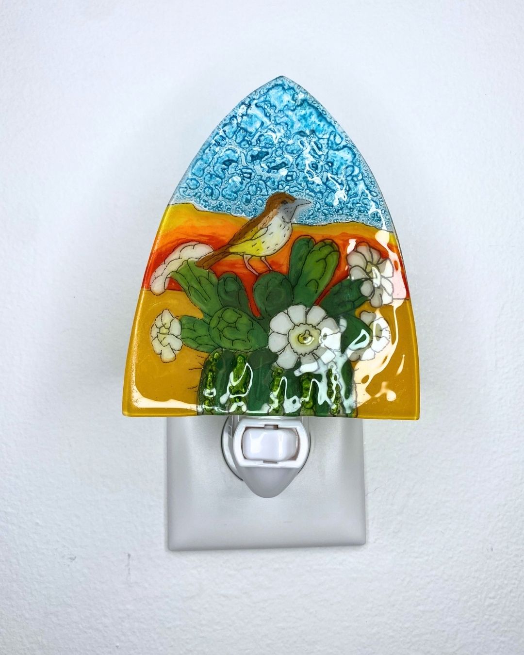 An unlit decorative glass night light plug-in featuring a colorful landscape design with a blue sky, desert background, blooming green cactus and a small bird, shown mounted on a white wall.