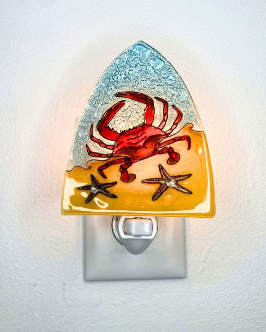 A decorative night light shaped like a beach scene, with a red crab on yellow sand, a blue textured sky, and two starfish, shown plugged into a wall outlet and glowing from behind.