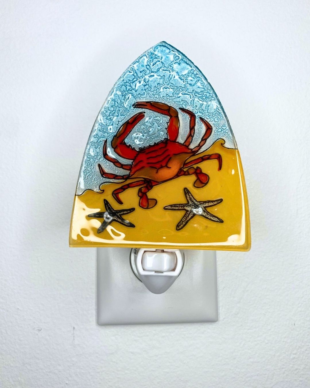A decorative glass night light shaped like a triangle, mounted on a white wall in a wall outlet. The design features a red crab on a yellow sandy beach with two starfish under a textured light blue ocean sky.