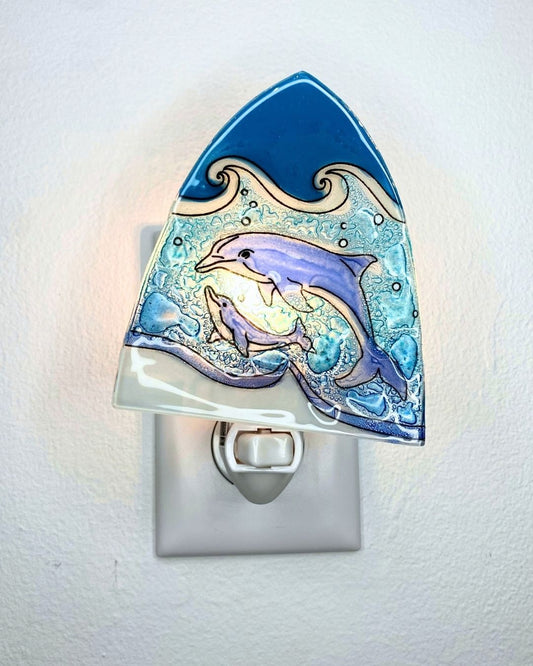 A glass night light shaped like a wave, featuring two dolphins swimming in blue  ocean waves. The night light is shown plugged into a white outlet against a white textured wall, emitting a warm glow.