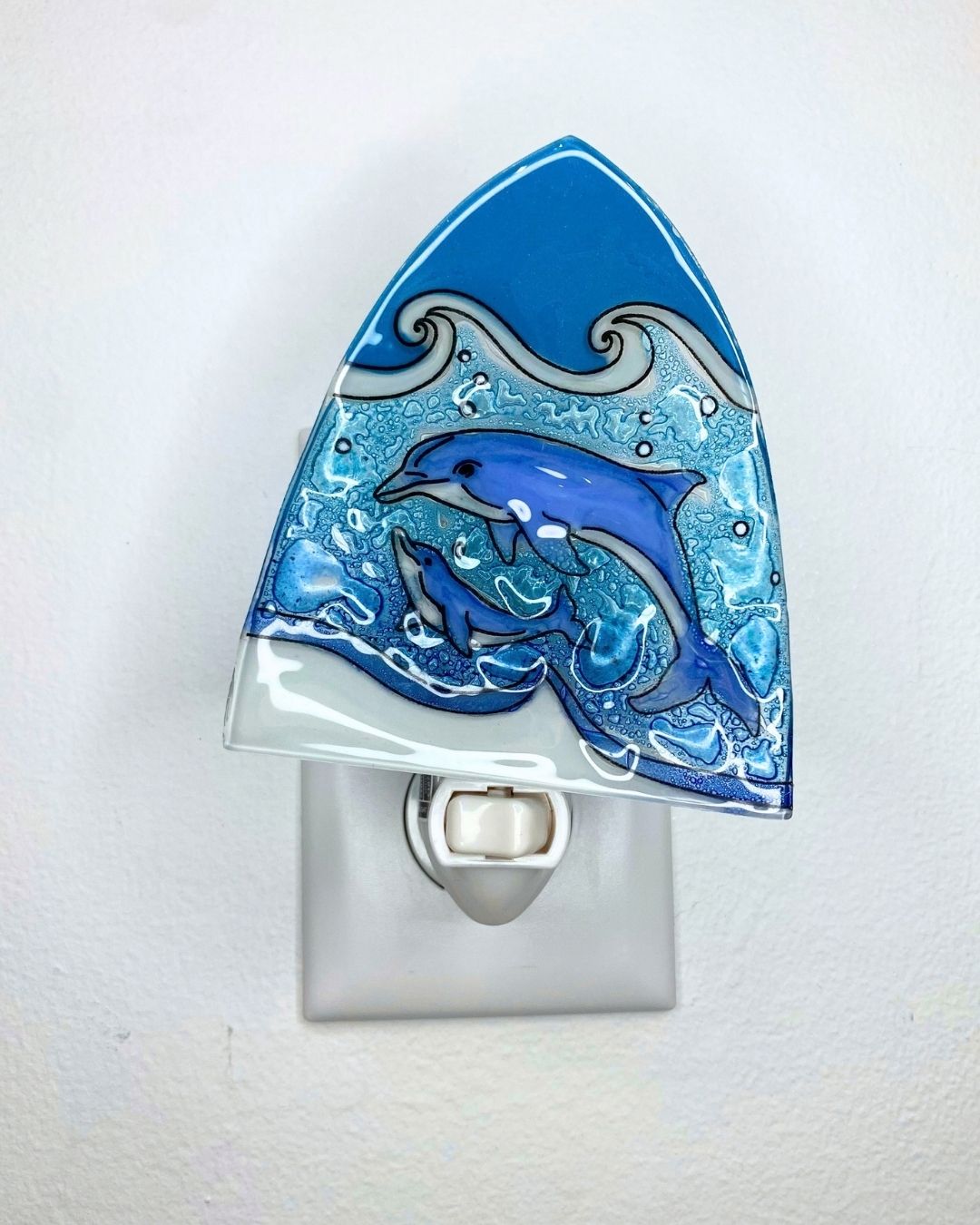 An unlit triangle-shaped blue dolphin night light with a wave design, shown plugged into a white wall outlet against a white wall.